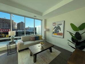A modern living room with a sofa, coffee table, and a large window offering a city view. - Spotless & Bright 1 BR downtown with private patio (Halifax)