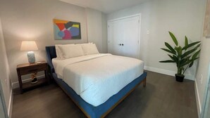 A bedroom with a bed, a nightstand, a lamp, a door, and a potted plant.