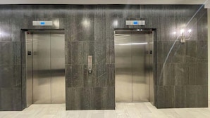 A modern elevator with metallic doors and a control panel, set in a hallway with dark tiled walls and light wooden flooring.