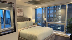 A modern bedroom with a large bed, a nightstand, a mirror, and a large window with a city view.