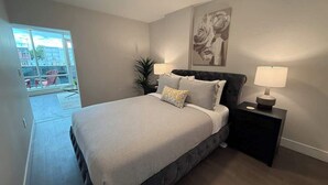 A bedroom with a large bed, a nightstand with a lamp, a potted plant, and a painting on the wall.