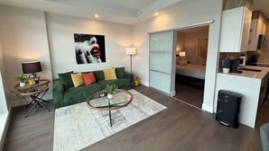 A modern living room with a green sofa, a coffee table, and a kitchen area visible through an open door.