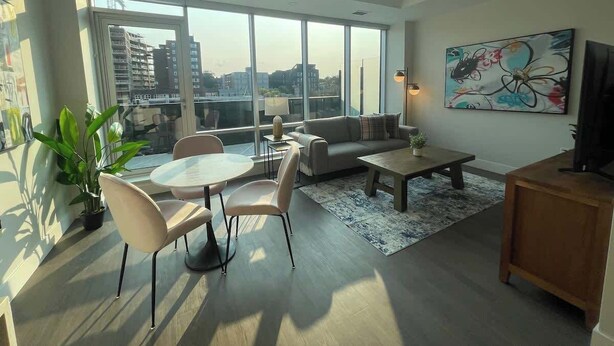 A modern living room with a round dining table, sofa, and coffee table. There is a large window with a view of buildings and a painting on the wall.