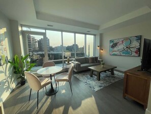 Ein modernes Wohnzimmer mit einem runden Esstisch, einer Couch und einem Couchtisch. Ein großes Fenster bietet Blick auf Gebäude, und an der Wand hängt ein Gemälde.