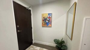 A hallway with a dark door, a patterned rug, a potted plant, and a framed painting on the wall.