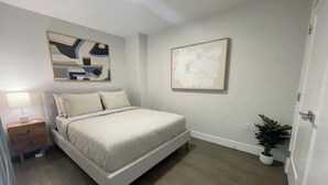 Ein Schlafzimmer mit Bett, Nachttisch, einem an der Wand hängenden abstrakten Gemälde, einer Topfpflanze und einer Tür.