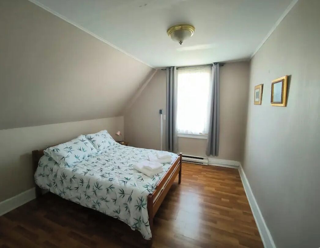 Een slaapkamer met een bed, een houten vloer, een plafondlamp, een raam met gordijnen en twee foto's in lijsten aan de muur.