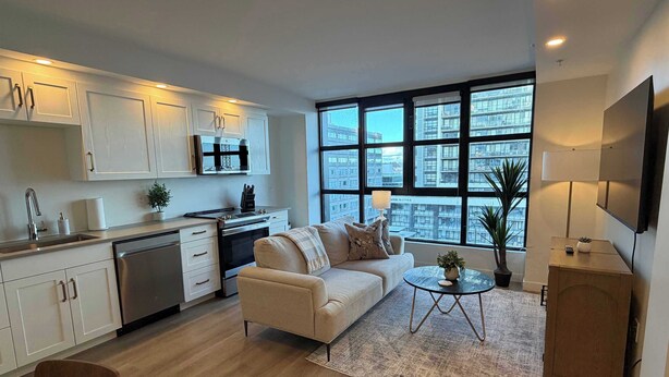 A modern kitchen with white cabinets, a stove, and a sink. There is a beige sofa and a small round table with a plant. The room has large windows with a city view.