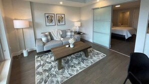 Una sala de estar moderna con un sofá, una mesa de centro y lámparas. Se ve una cama a través de una puerta abierta.
