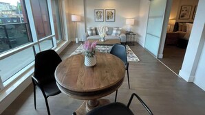 Una sala de estar moderna con una mesa redonda de madera, dos sillas negras, un sofá y una mesa de centro con un jarrón de flores. Hay dos lámparas de pared y obras de arte enmarcadas sobre el sofá.