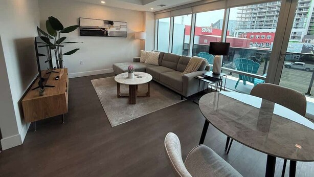 A modern living room with a sofa, coffee table, and dining table. There is a plant, a lamp, and a view of a building outside. - Bright, spotless and modern 1 BR with gym (Halifax)