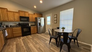 A kitchen with wooden cabinets, a stainless steel refrigerator, a microwave, and a dining area with a table and chairs. - Family-friendly 3BR with a fun 90s themed game room, fenced yard & grill (Broken Arrow)