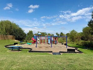 A playground with slides and climbing structures, surrounded by grass and trees.