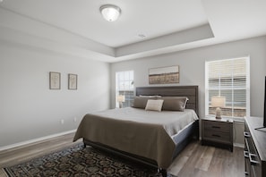 A bedroom with a bed, bedside tables, a nightstand, a lamp, a rug, and a window with blinds. - Family-friendly 3BR home with a fun game room, fenced yard, patio, and extra sleeping space—perfect for groups and longer stays. (Broken Arrow)