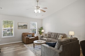 A living room with a ceiling fan, two windows with blinds, a brown leather chair, a grey sofa, a small round table, and a floor lamp.
