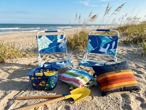 Strandstole, en strandtaske, et håndklæde, en strandpude og en skovl ligger i sandet.
