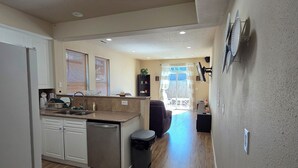 A modern kitchen with a dishwasher, a sink, and a window with blinds. There is a dining area with a table and chairs, and a living room with a sofa.