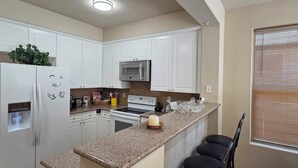 A kitchen with white cabinets, a granite countertop, a microwave, a stove, and a refrigerator. There are three bar stools and a window with blinds.