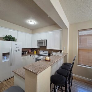 A kitchen with white cabinets, a granite countertop, a microwave, a stove, and a refrigerator. There are three bar stools and a window with blinds.