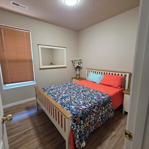 A bedroom with a wooden bed, a nightstand, a mirror, a window with blinds, and a vase with flowers. - Family-Friendly One-Level Home in Aloha (Beaverton)