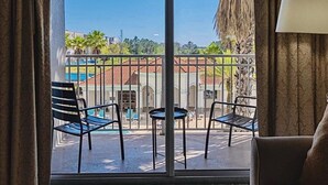 Ein Balkon mit Metallstühlen und einem kleinen Tisch, der auf einen Innenhof mit einem Gebäude mit Ziegeldach und Palmen blickt.