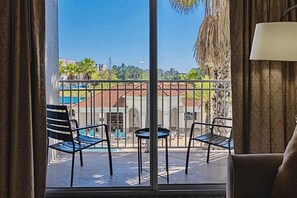 A balcony with metal chairs and a small table, overlooking a courtyard with a tiled-roof building and palm trees.