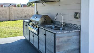 Cocina exterior con parrilla integrada y pileta, frente a una pared de casa con fachada de piedra.