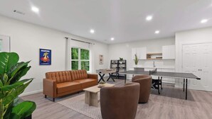 Uma sala de estar moderna com um sofá de couro marrom, duas poltronas marrons, uma mesa de centro e uma mesa de pingue-pongue. Há uma área de cozinha com armários brancos e uma janela com vista para o verde.