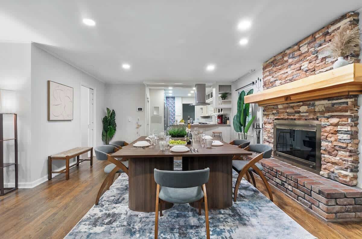 A modern dining area with a wooden table and chairs, a stone fireplace, and a view into the kitchen.