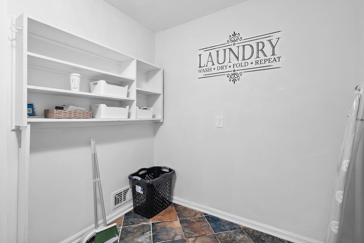 Una sala de bugaderia amb prestatges blancs, una cistella de roba negra i un cartell a la paret que diu "LAUNDRY WASH DRY FOLD REPEAT".