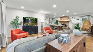 Un salon moderne avec un canapé gris, un fauteuil rouge et une table basse en bois. Un téléviseur est fixé au mur, et on aperçoit un coin cuisine au fond de la pièce.