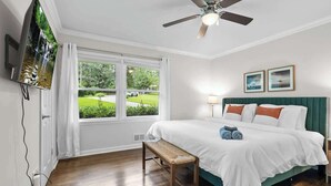 Une chambre à coucher comprenant un lit, un banc, un ventilateur de plafond, une télévision et une fenêtre donnant sur un paysage verdoyant.