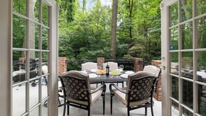 Une terrasse ouverte avec une table dressée pour quatre personnes, entourée de piliers en briques, et une vue sur une zone boisée à travers de grandes portes vitrées.
