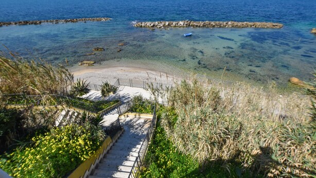 Un percorso costiero che conduce a una spiaggia con acque limpide e affioramenti rocciosi.