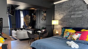 A modern bedroom with a large bed, a small desk, a chair, a sofa, a wall-mounted TV, a lamp, and a decorative wall. - La 104 private cocooning rooms with whirlpool bath (Saint-Omer)
