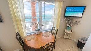 A dining area with a wooden table, chairs, a small potted plant, a television on the wall, and a view of the ocean through the window. - Beachside Cottage #3/ Fun In The Sun/Dogs Welcome (Redington Shores)