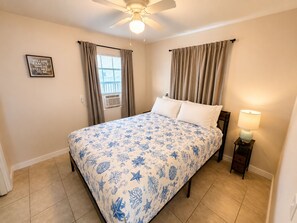 A bedroom with a bed, curtains, a ceiling fan, a bedside lamp, and a framed quote on the wall.