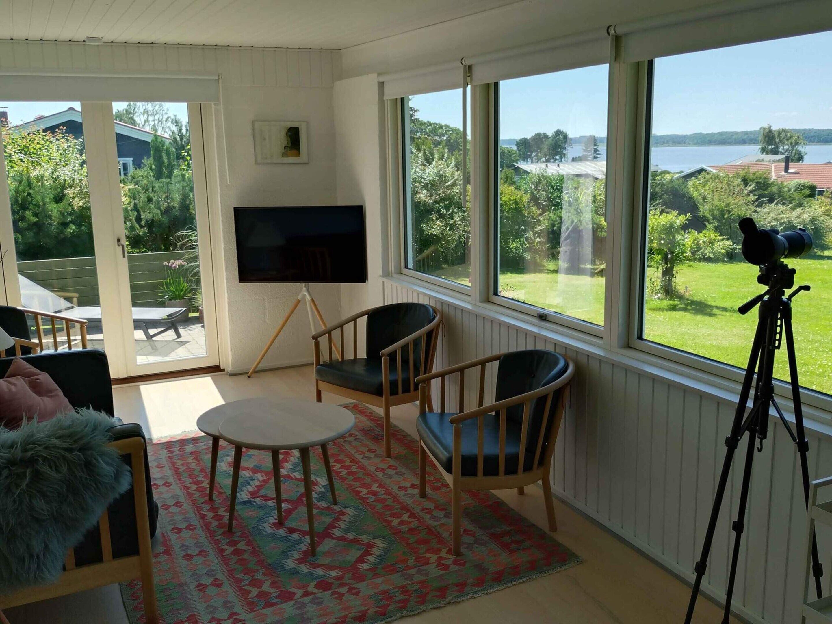A room with a large window, a television, two chairs, a small table, and a telescope on a tripod.