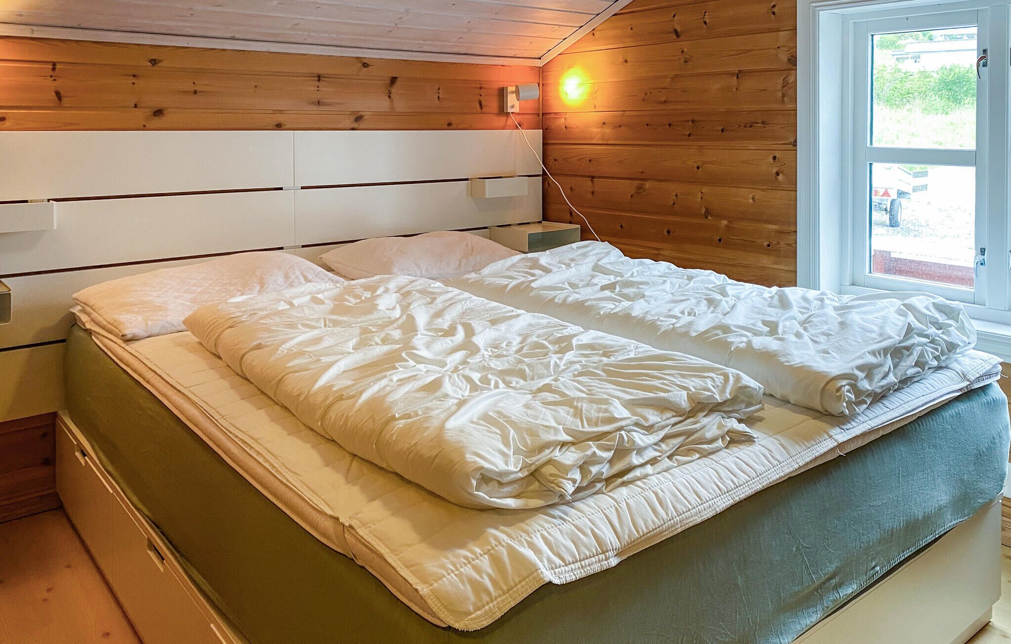 Un lit avec du linge de lit blanc, dans une pièce aux murs en bois et dotée d’une fenêtre.