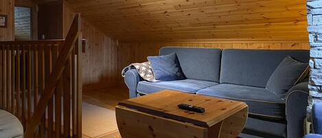 Een gezellig chaletinterieur met een houten plafond, een blauwe bank met kussens en een kleine houten tafel met een afstandsbediening.