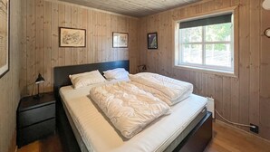 Ein Schlafzimmer mit Holzboden und -wänden, einem Bett mit weißer Bettwäsche, zwei gerahmten Bildern an der Wand und einem Fenster mit Blick ins Grüne.