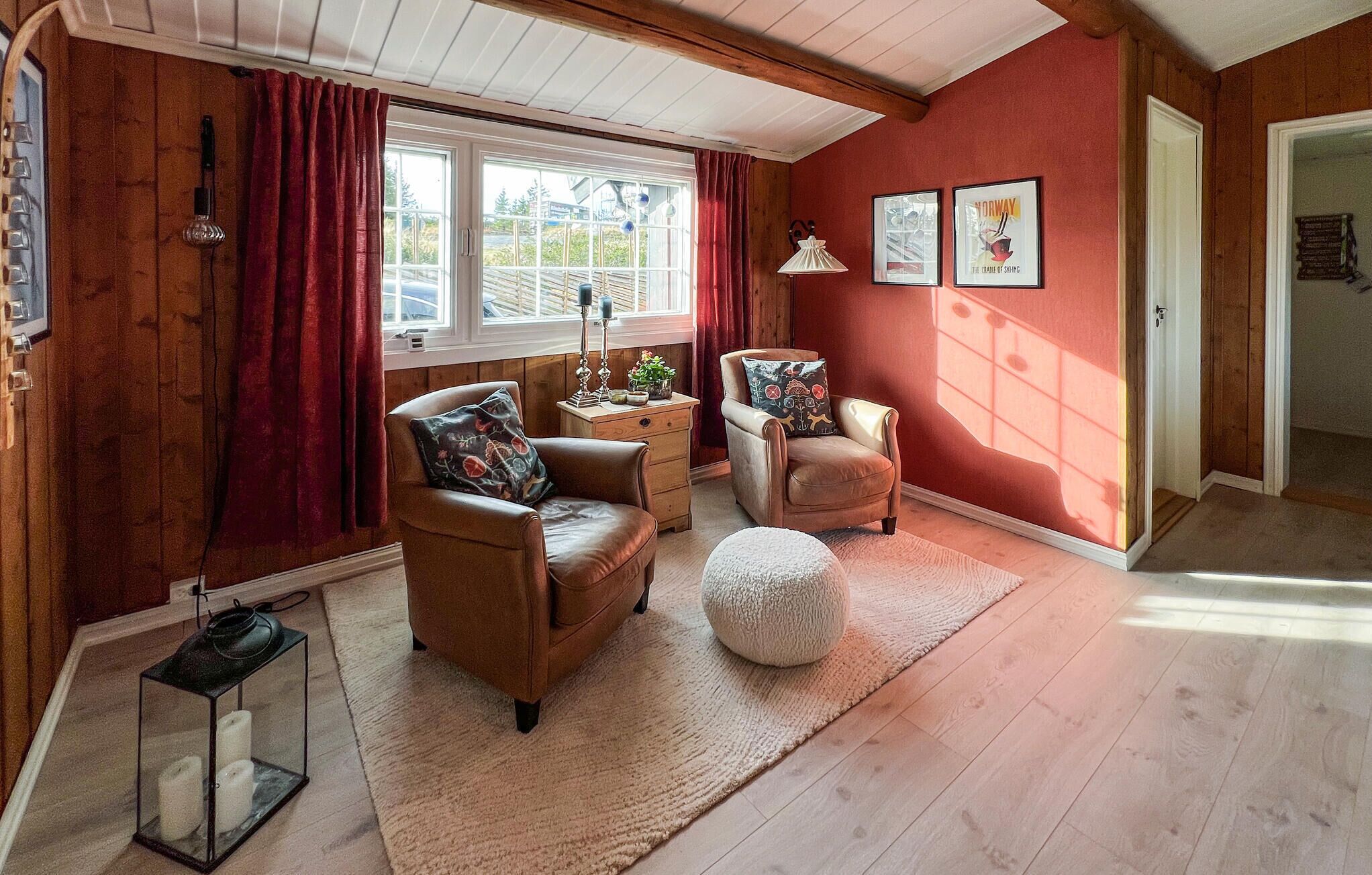 A cozy living room with wooden floors, a red wall, and a large window with curtains. There are two leather armchairs, a small table with a lamp, and a white pouf.