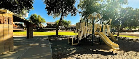 A playground with a slide and climbing structures, surrounded by trees and a paved walkway.