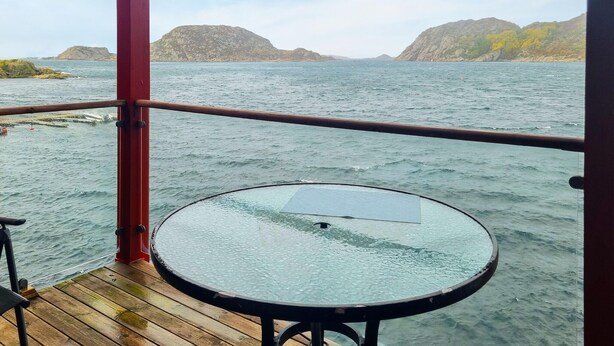 Une table ronde en verre, posée sur une terrasse en bois, offre une vue sur la mer et les collines rocheuses qui se dessinent à l’horizon.