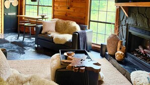 L’intérieur chaleureux d’un chalet avec une cheminée, un téléviseur fixé au mur, un coin repas avec une table et des chaises, et une grande fenêtre donnant sur le paysage extérieur.