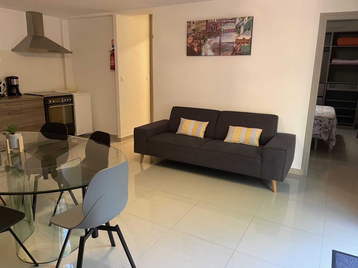 A modern living room with a glass dining table, a sofa, and a kitchen area with a stove and cabinets.