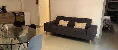 A modern living room with a glass dining table, a sofa, and a kitchen area with a stove and cabinets.