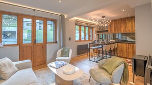A spacious living room with a sofa, armchair, and coffee table. A kitchen area with wooden cabinets and a dining table is visible in the background. - Duplex Veyra - Welkeys (Megève)