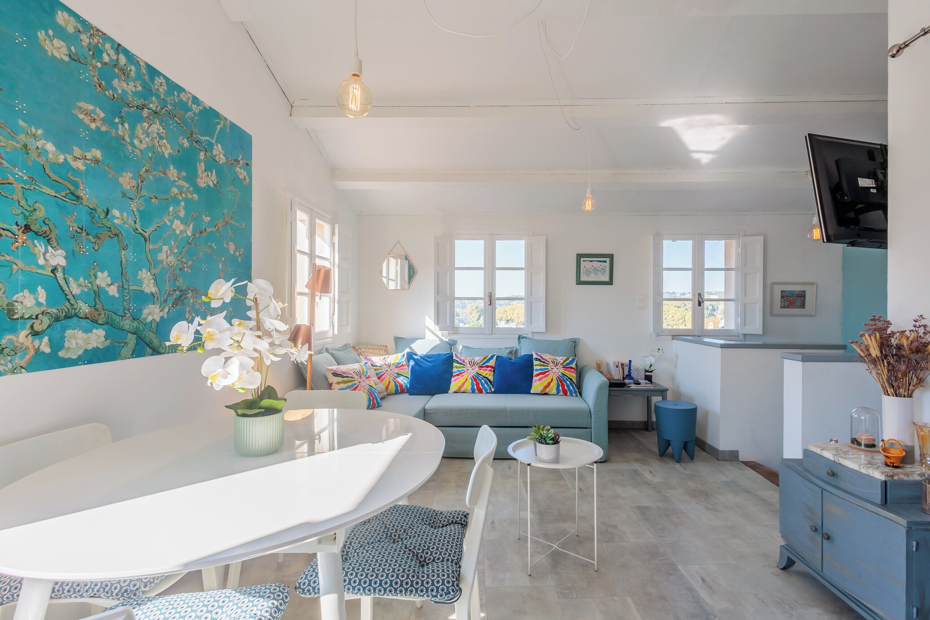 Un salón luminoso con una mesa blanca, sofá azul y un cuadro grande con motivos florales.