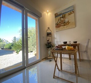Une pièce dotée d’une grande porte-fenêtre coulissante donnant sur un jardin, d’un tableau accroché au mur, d’une table avec des fruits et des bols, et d’une chaise.